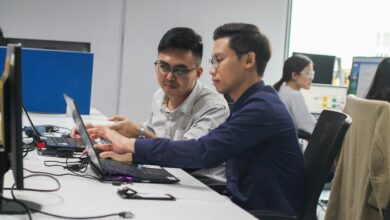 Two digital marketers collaborating on a project in a shared office space, surrounded by colleagues working on laptops and marketing dashboards.